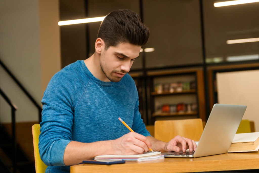 Joven opositor estudiando con portátil y cuaderno en biblioteca, preparándose para el examen de la Guardia Civil